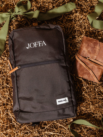 Flat lay of JOFFA-branded backpack and leather wrap journal placed on kraft crinkle paper with green ribbon, styled as an elevated and meaningful corporate gift with impact-driven design.