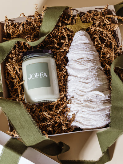 Flat lay of a JOFFA candle and white washcloth tree with gold star, packed in eco-friendly crinkle paper inside a white gift box, accented with green ribbon for corporate holiday gifting.