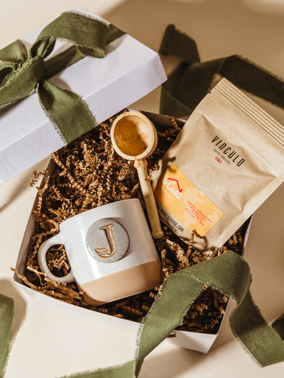 Flat lay of a Su Casa speciality coffee sample, “J” ceramic mug, and wooden scoop nestled in crinkle paper inside a white gift box with an olive green ribbon—styled for meaningful corporate gifting.
