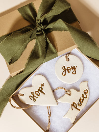 Gift box opened to reveal ceramic “Hope,” “Peace,” and “Joy” ornaments nestled inside, accented with a frayed green ribbon for a heartfelt and meaningful holiday gifting presentation.