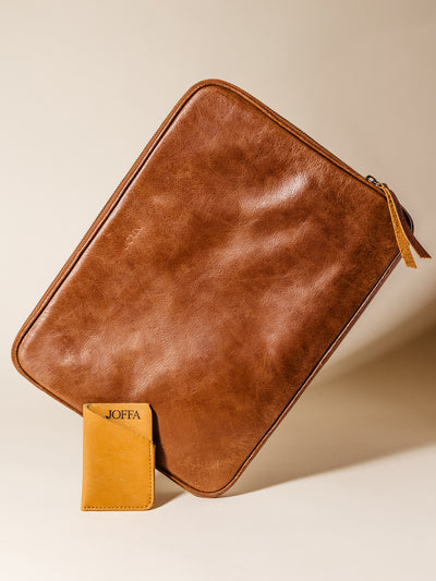 Brown leather tech sleeve propped upright with a mustard yellow card holder leaning against it, featuring clean stitching and debossed JOFFA branding, styled against a soft beige background.