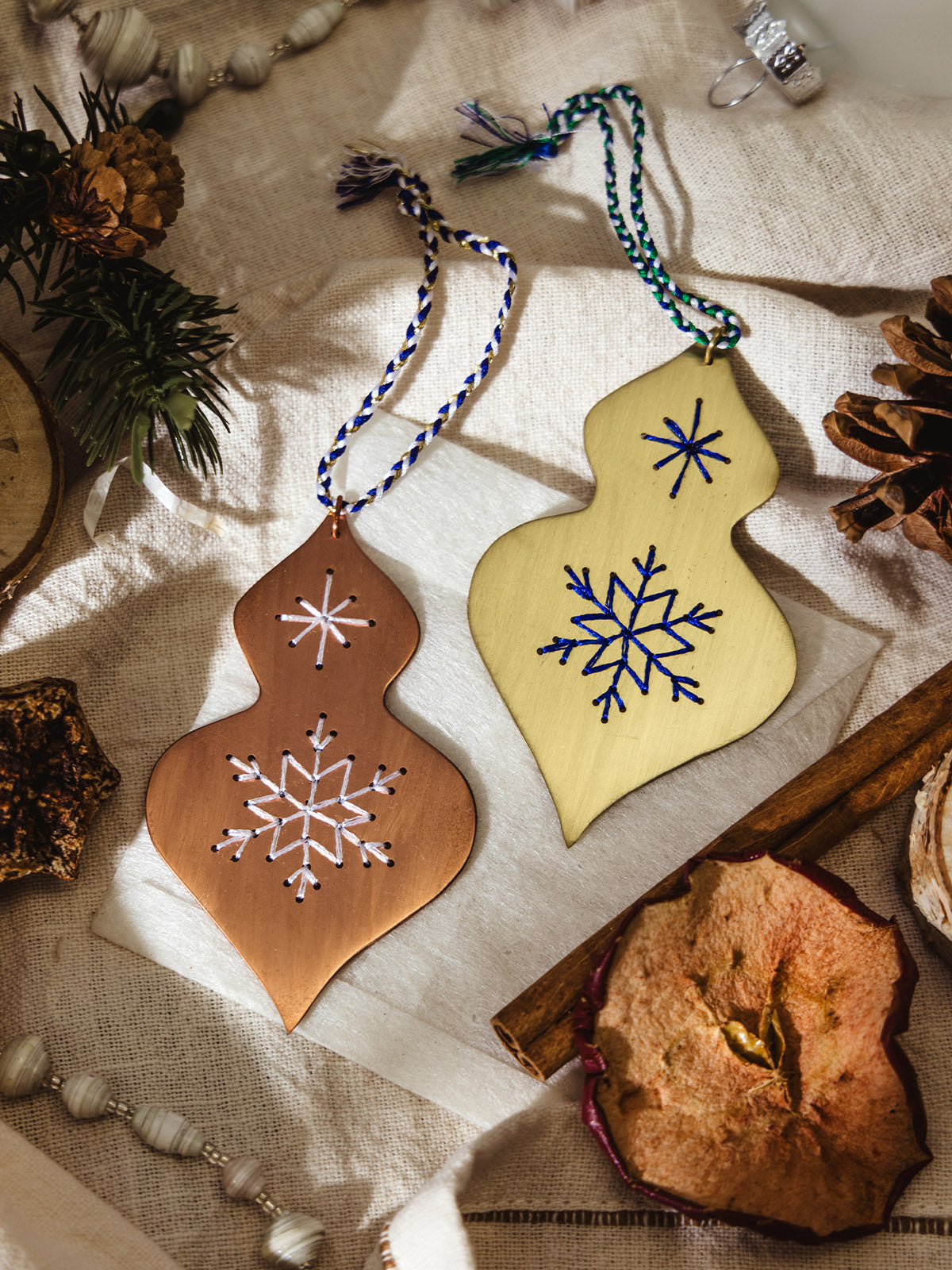 Two handmade holiday ornaments, one copper and one brass, each embroidered with stitched snowflakes, lying on a linen cloth surrounded by pinecones, dried fruit, and festive greenery.