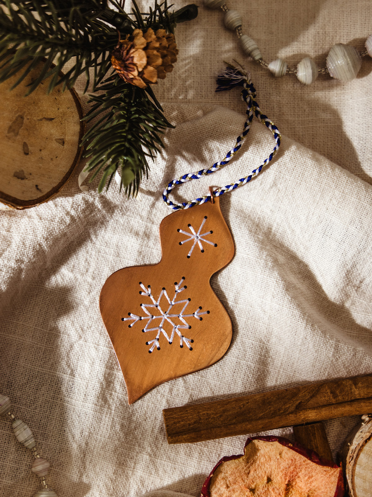 Close-up of a copper ornament embroidered with white stitched snowflakes, displayed on a neutral linen background with pine branches and cinnamon sticks.