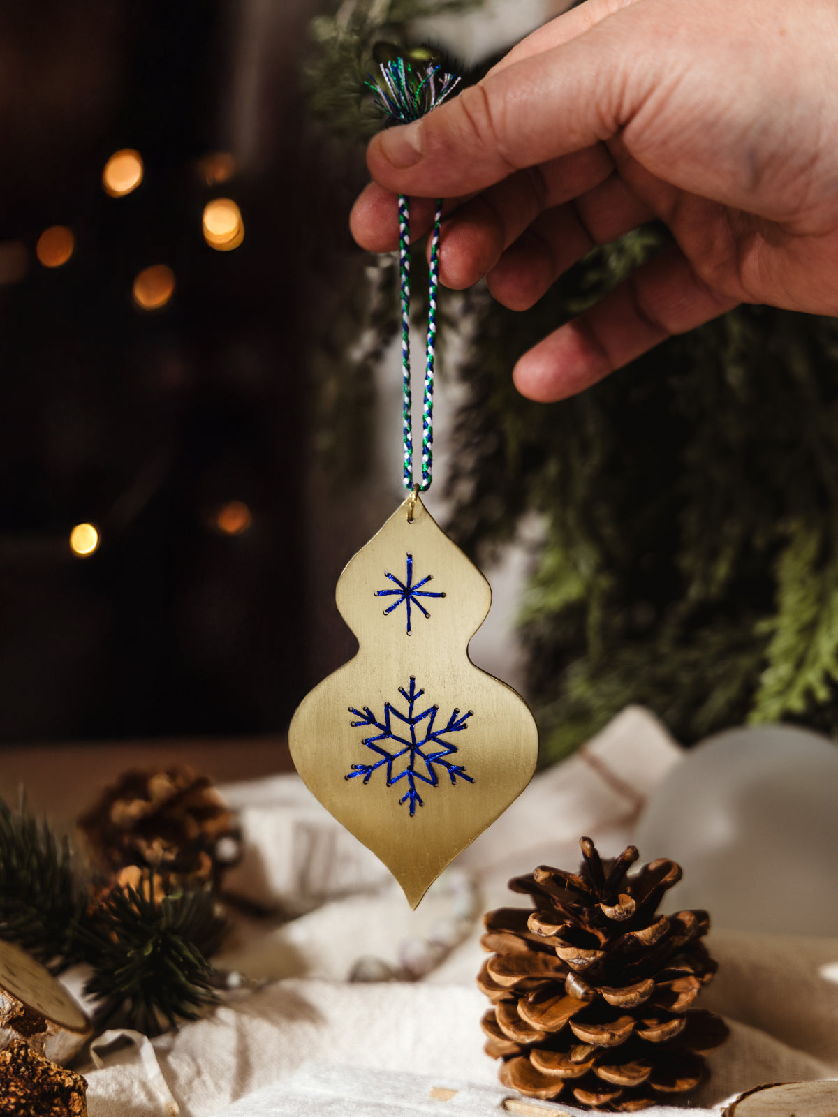 Hand holding a brass ornament with blue stitched snowflakes, ready to hang against a backdrop of pine branches and warm holiday lights.