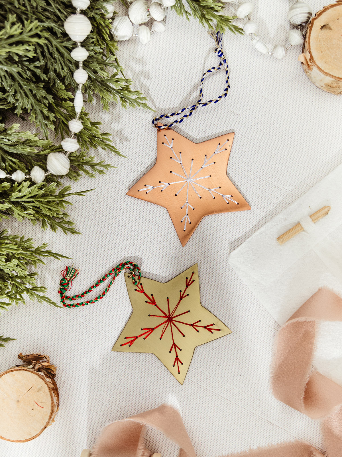 A pair of star-shaped ornaments, one brass with red embroidered snowflake and one copper with white embroidered snowflake, displayed among pine garland and ribbon.