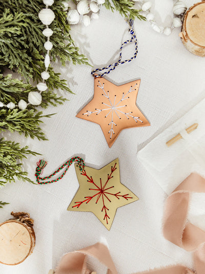 A pair of star-shaped ornaments, one brass with red embroidered snowflake and one copper with white embroidered snowflake, displayed among pine garland and ribbon.