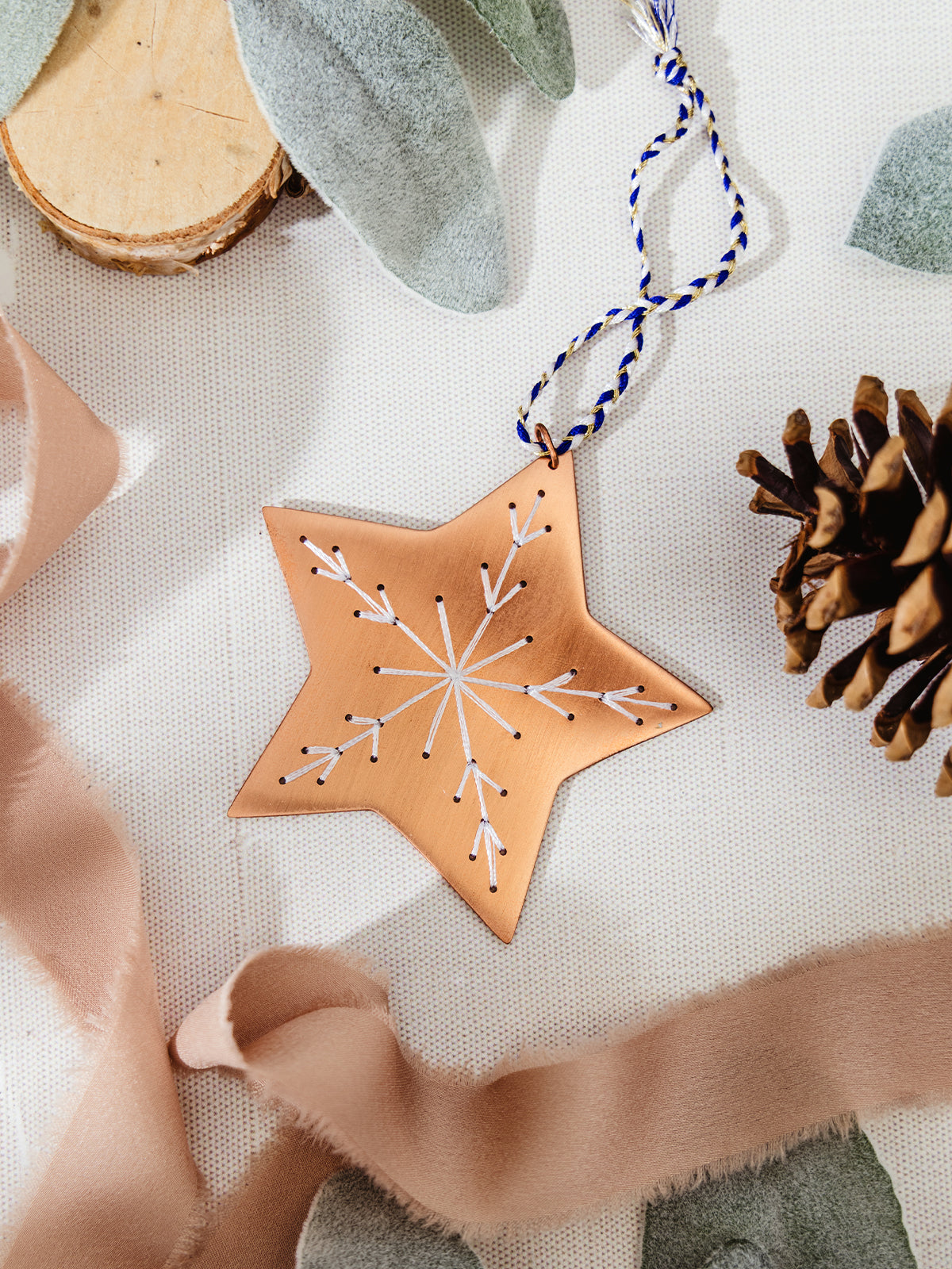 Copper star ornament with white embroidered snowflake, displayed on a linen surface with ribbon, pinecone, and soft felt leaves.