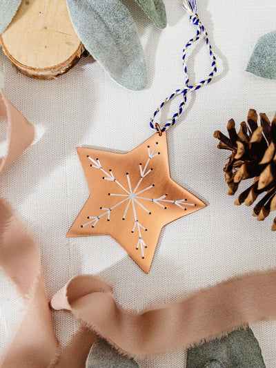 Copper star ornament with white embroidered snowflake, displayed on a linen surface with ribbon, pinecone, and soft felt leaves.