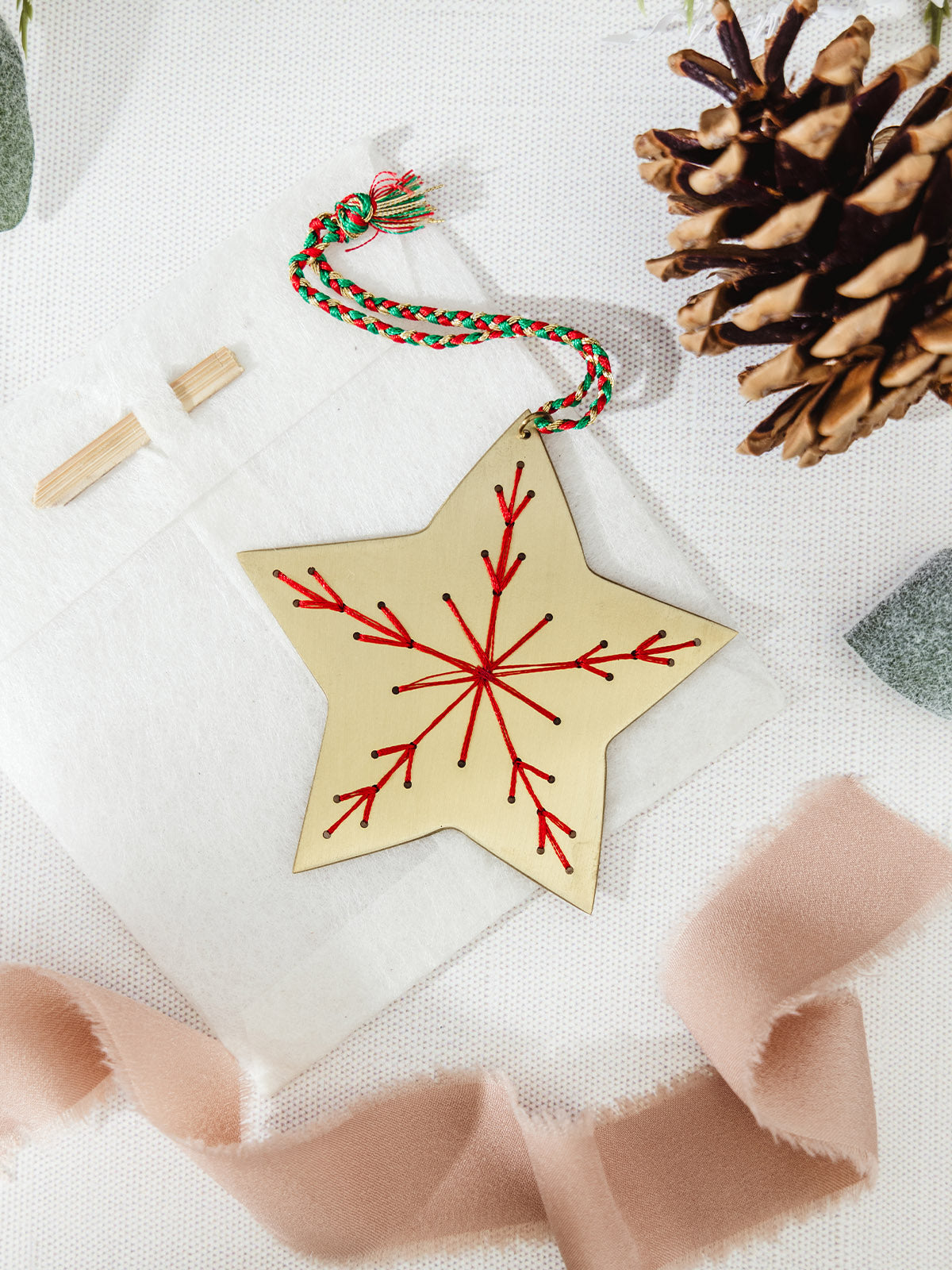 Brass star ornament with bold red embroidered snowflake, lying on a linen background beside ribbon and a pinecone.