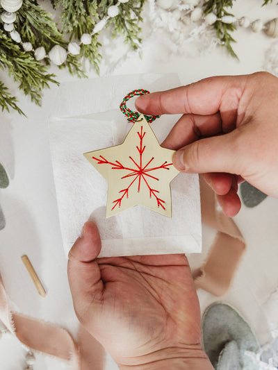 Hands holding a brass star ornament with a red stitched snowflake, above a linen pouch with holiday greenery in the background.