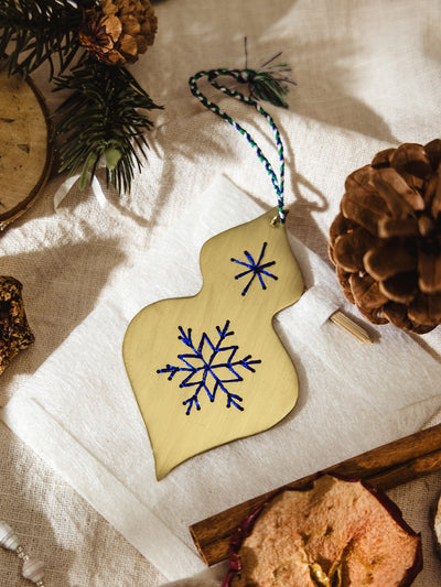 Close-up of a brass ornament embroidered with blue stitched snowflakes, displayed on a neutral linen background with pine branches and cinnamon sticks.