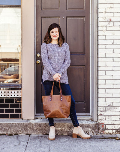 Woman standing outside with a handcrafted brown leather tote bag from Beloved Leather, styled with a cozy sweater, dark jeans, and ankle boots — ethical and timeless everyday bag.