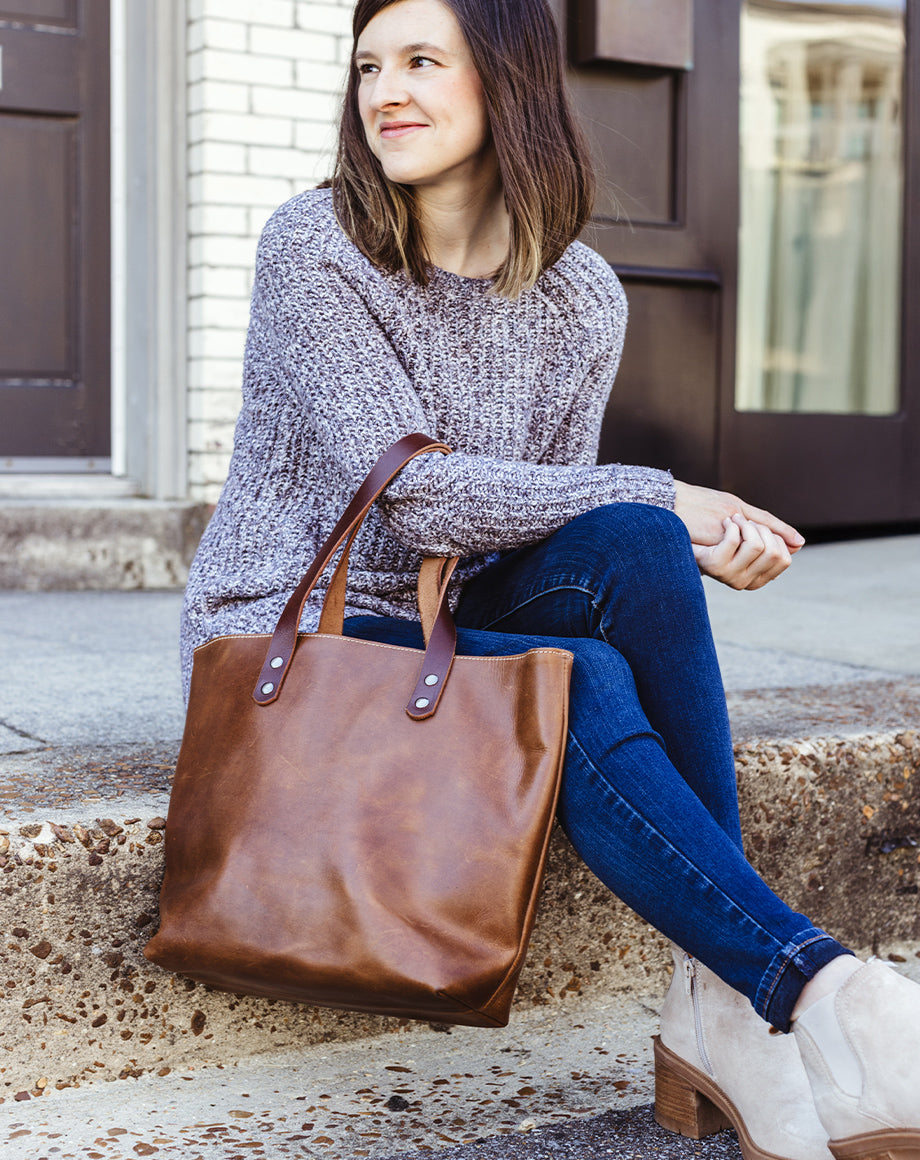 Close-up of woman sitting on outdoor steps holding a Beloved Leather brown tote bag, styled with casual jeans, a cozy sweater, and neutral suede ankle boots — sustainably made leather handbag.