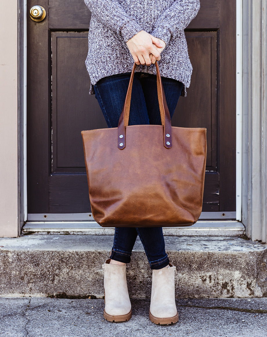 Front view of artisan-made brown leather tote bag from Beloved Leather held by a woman wearing jeans and ankle boots, showcasing durable minimalist design for everyday use.