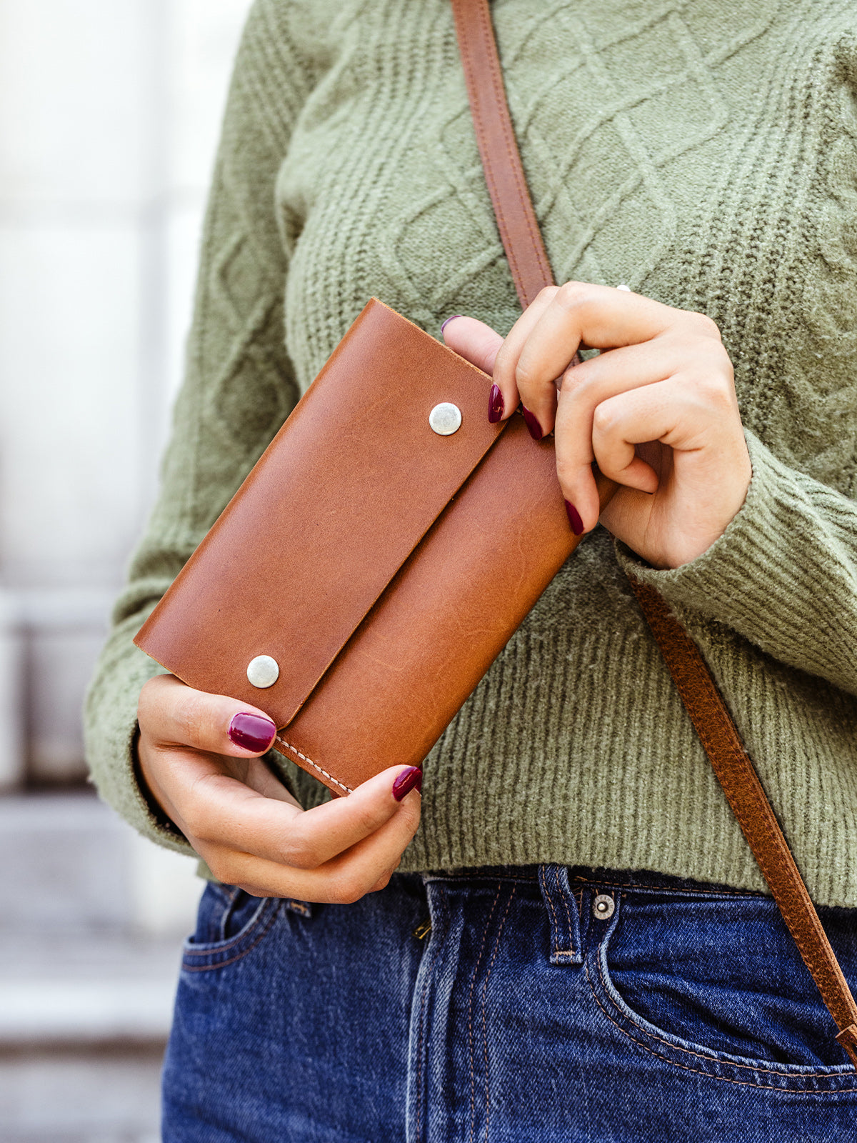 Woman holding a handcrafted tan leather wallet with silver snap closure, styled with a green sweater and crossbody bag — ethically made minimalist accessory.
