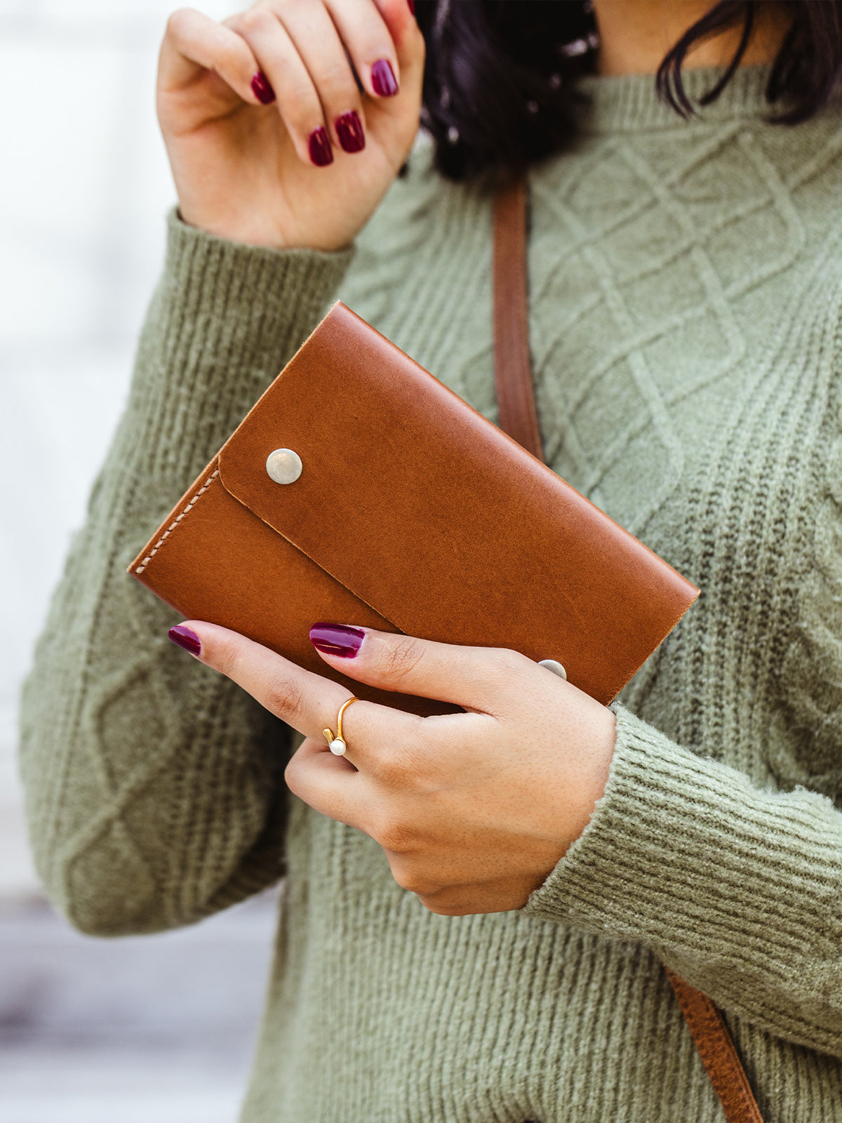 Close-up of hands showing a tan leather wallet with silver snaps, paired with a green sweater and gold ring — fair trade handmade leather wallet.