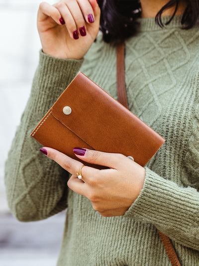 Close-up of hands showing a tan leather wallet with silver snaps, paired with a green sweater and gold ring — fair trade handmade leather wallet.