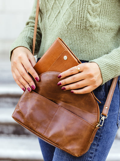 Woman placing a tan leather wallet into a matching brown leather crossbody bag while wearing a casual green sweater and jeans — sustainably made leather goods.