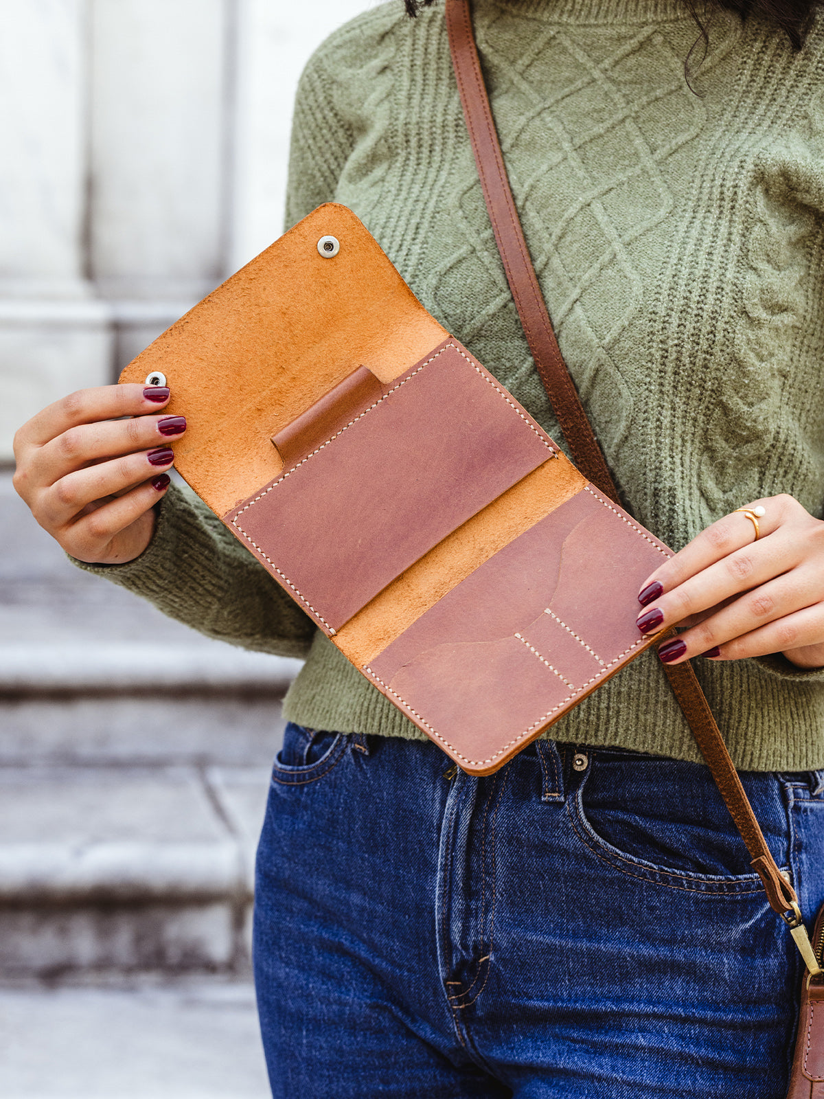 Woman holding open a tan leather wallet to show interior card slots and cash pocket, paired with a green sweater and jeans — artisan-made sustainable leather accessory.