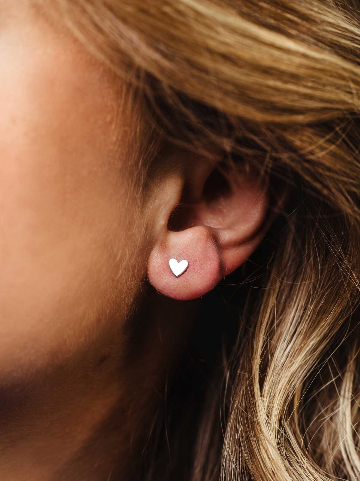 Close-up of a woman wearing small silver heart stud earrings, highlighting a simple and elegant design that pairs effortlessly with everyday style.