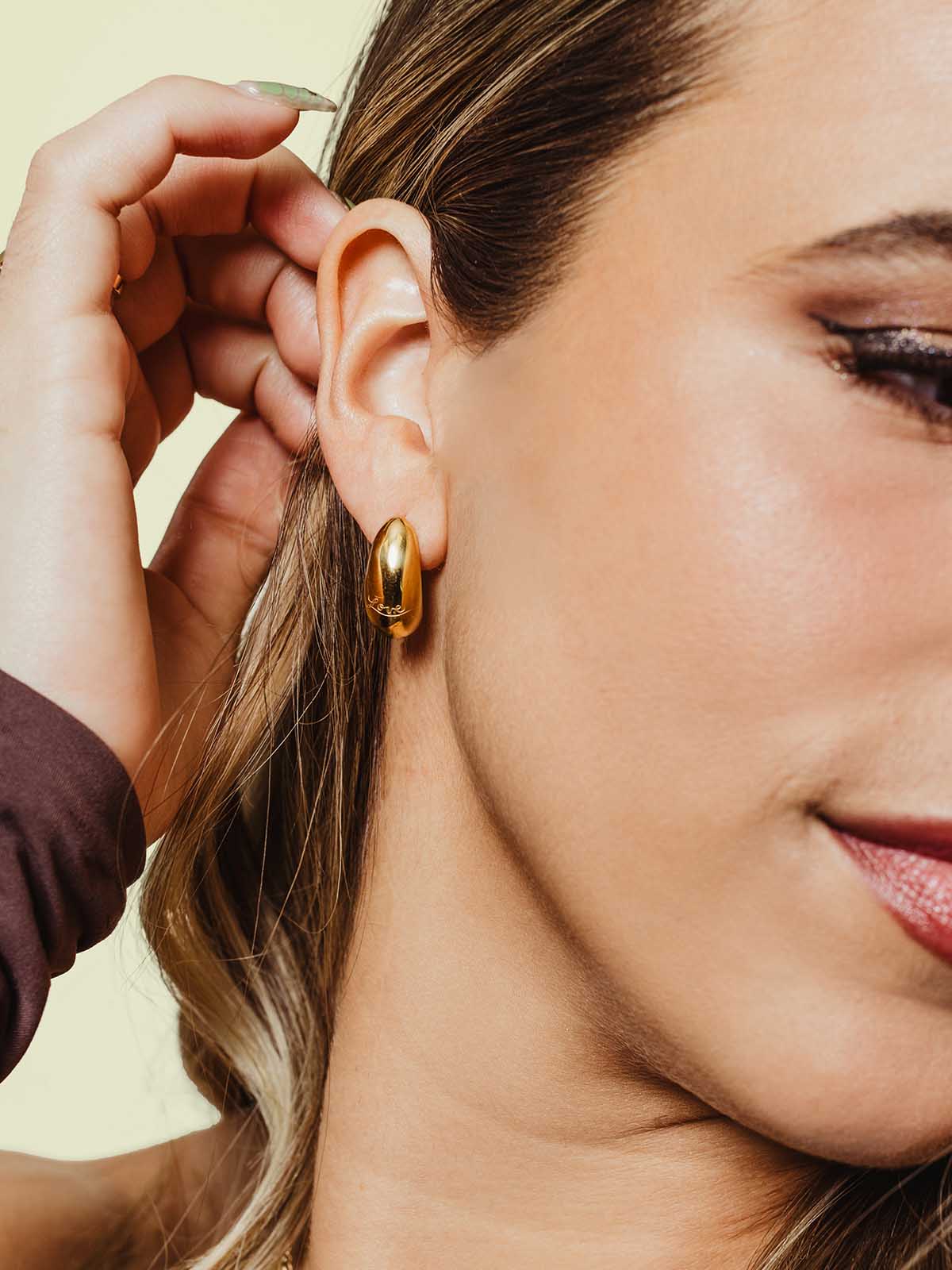 Close-up of a woman wearing bold gold hoop earrings with a smooth, polished finish. Her hair is tucked behind her ear as she gently touches it, highlighting the handcrafted shine of artisan-made jewelry.