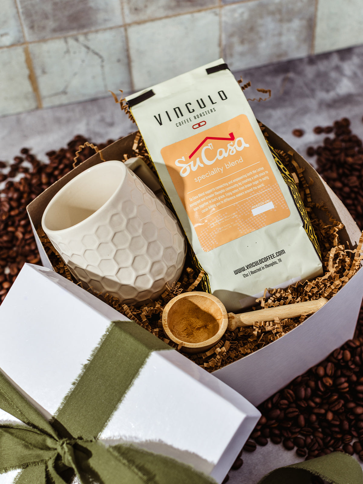 Vinculo Coffee Roasters Su Casa specialty blend gift set with white honeycomb ceramic mug, wooden coffee scoop, and festive packaging on coffee beans background.