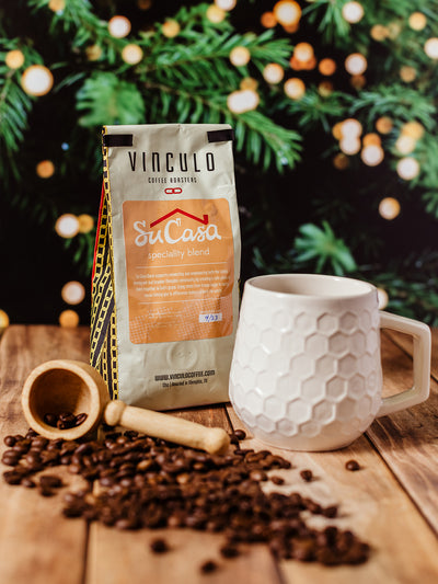 Vinculo Coffee Roasters Su Casa specialty blend with white honeycomb ceramic mug and wooden scoop surrounded by roasted coffee beans on rustic wood table, Christmas lights in background.