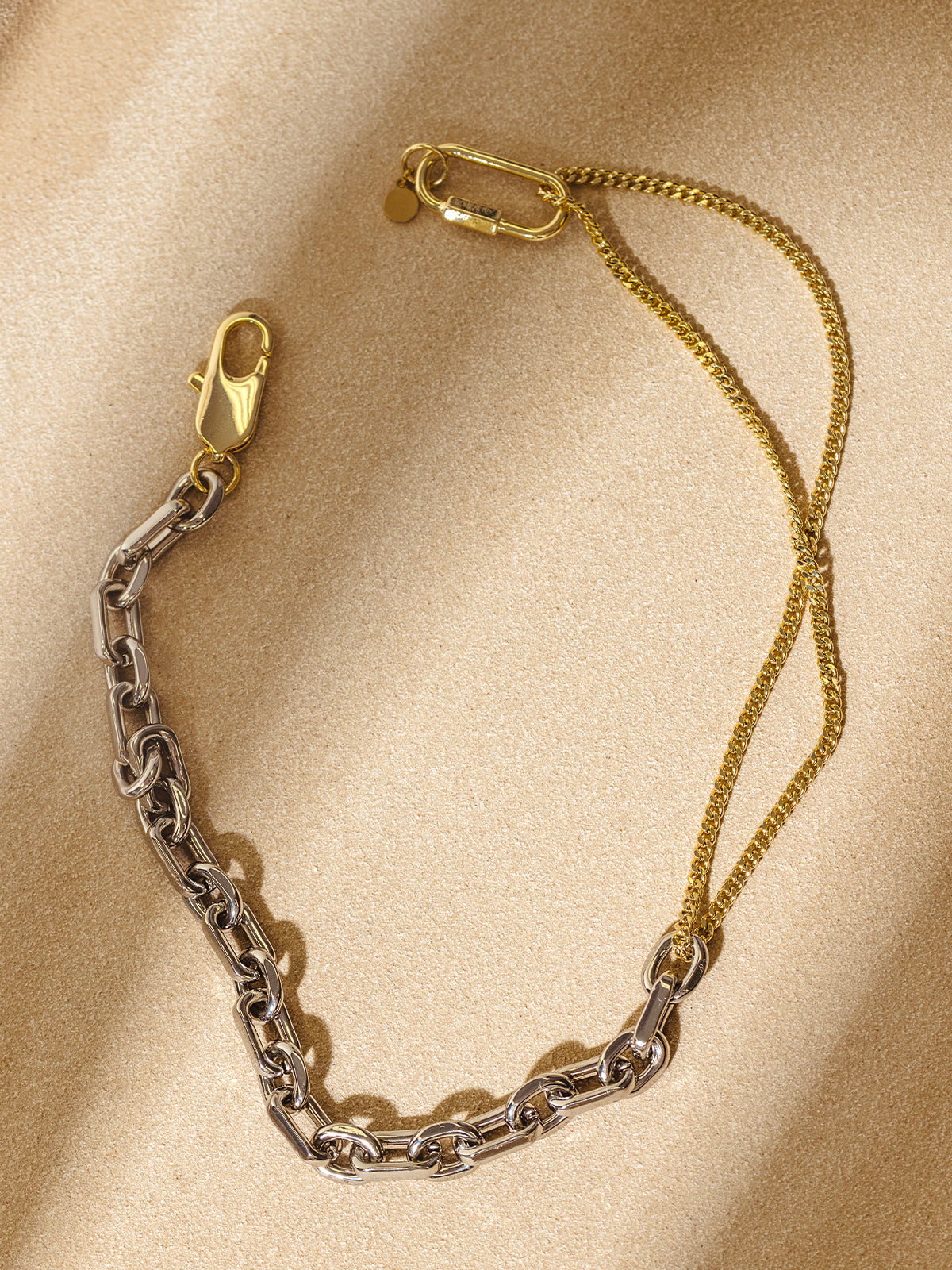 A two-tone chain necklace with silver and gold links displayed on a warm sand-colored background. The layered gold chain adds a delicate contrast to the bold silver links, creating a striking, contemporary style.