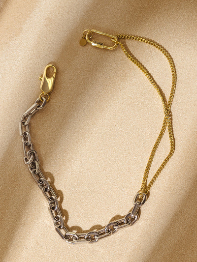 A two-tone chain necklace with silver and gold links displayed on a warm sand-colored background. The layered gold chain adds a delicate contrast to the bold silver links, creating a striking, contemporary style.
