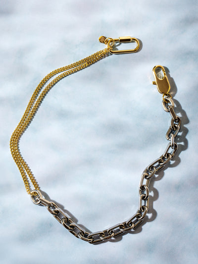 A two-tone chain necklace featuring a mix of silver and gold links, laid out on a soft blue background. The necklace includes a gold carabiner clasp and a delicate gold chain accent, creating a modern and edgy design.