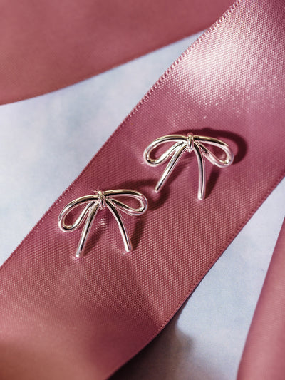 A pair of silver bow stud earrings displayed on a deep mauve satin ribbon. The polished silver finish gleams under soft lighting, showcasing the delicate and feminine design.