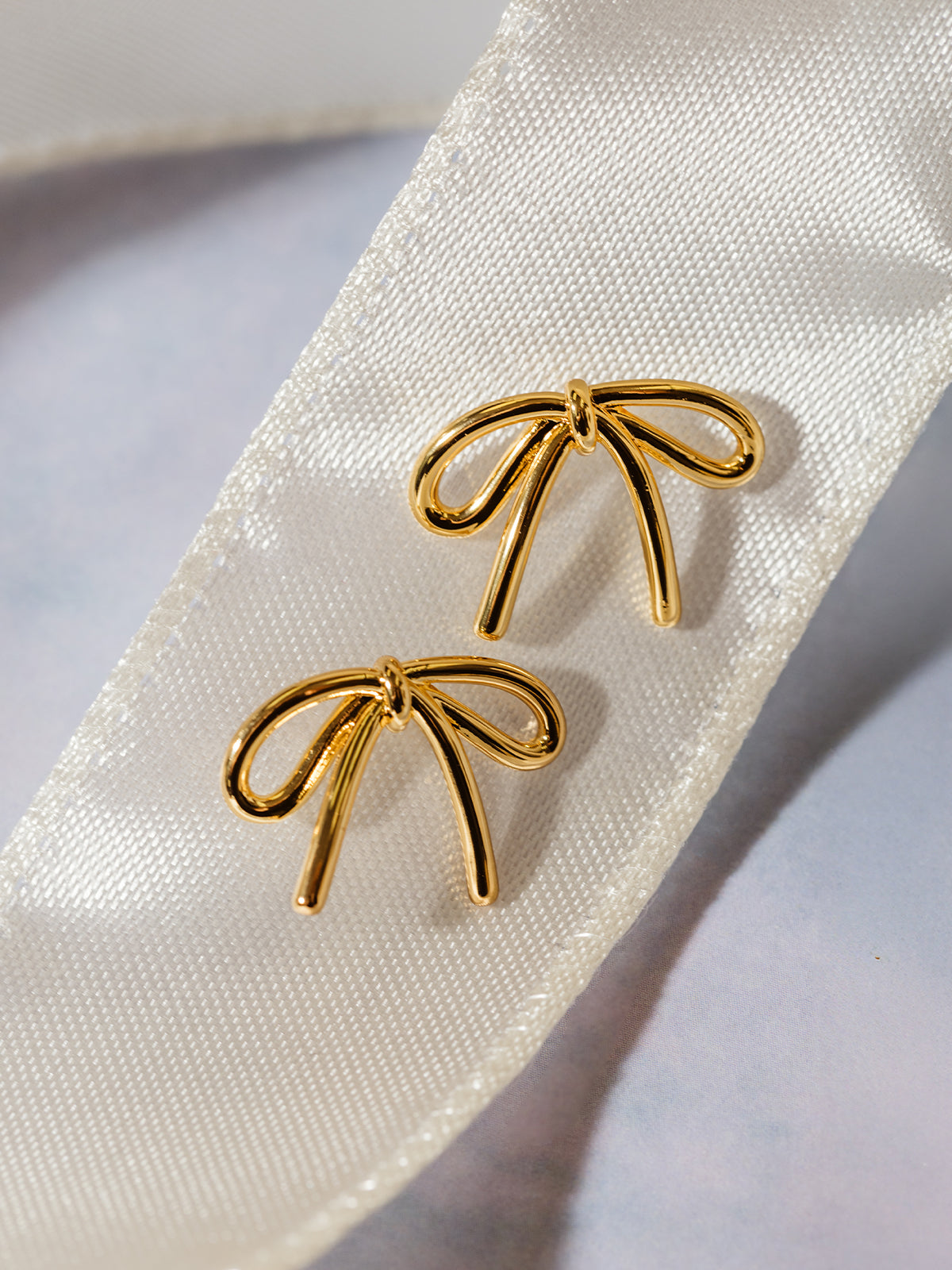 A pair of gold bow stud earrings elegantly placed on a cream satin ribbon. The warm gold tone contrasts beautifully against the soft fabric, emphasizing the dainty and timeless style.