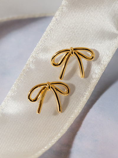 A pair of gold bow stud earrings elegantly placed on a cream satin ribbon. The warm gold tone contrasts beautifully against the soft fabric, emphasizing the dainty and timeless style.