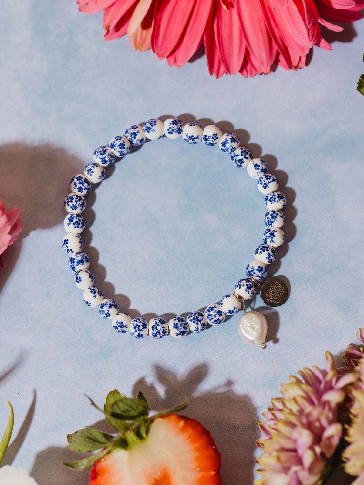 Alt text:
"Stretch bracelet made of round white beads with blue floral patterns, featuring a pearl charm and metallic tag, displayed flat on a pastel background with flowers and a sliced strawberry.