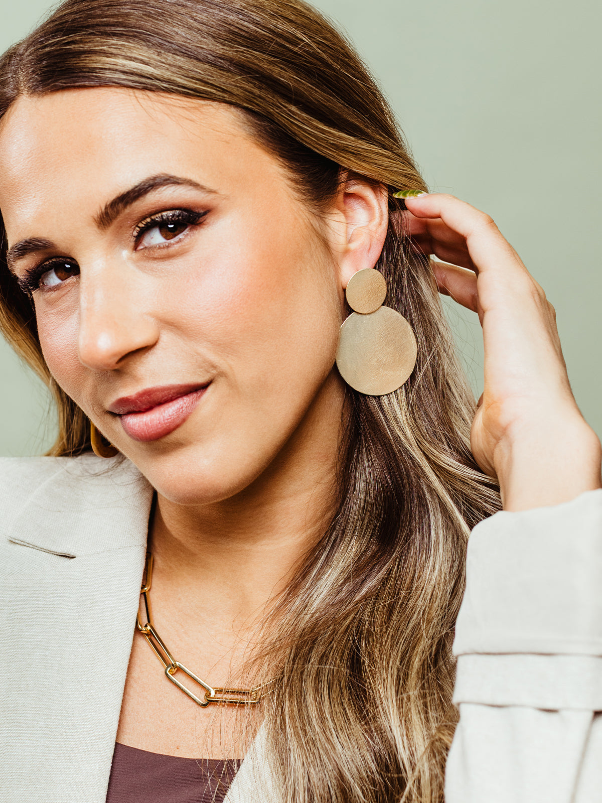  A stylish woman wearing oversized gold statement earrings and a chunky gold chain necklace. She gently tucks her hair behind her ear, showing off the bold, textured design of the earrings.