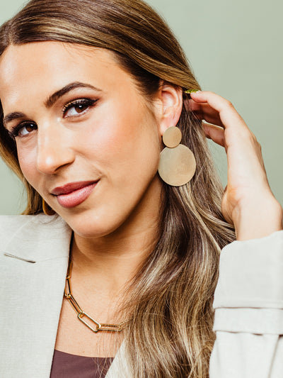  A stylish woman wearing oversized gold statement earrings and a chunky gold chain necklace. She gently tucks her hair behind her ear, showing off the bold, textured design of the earrings.