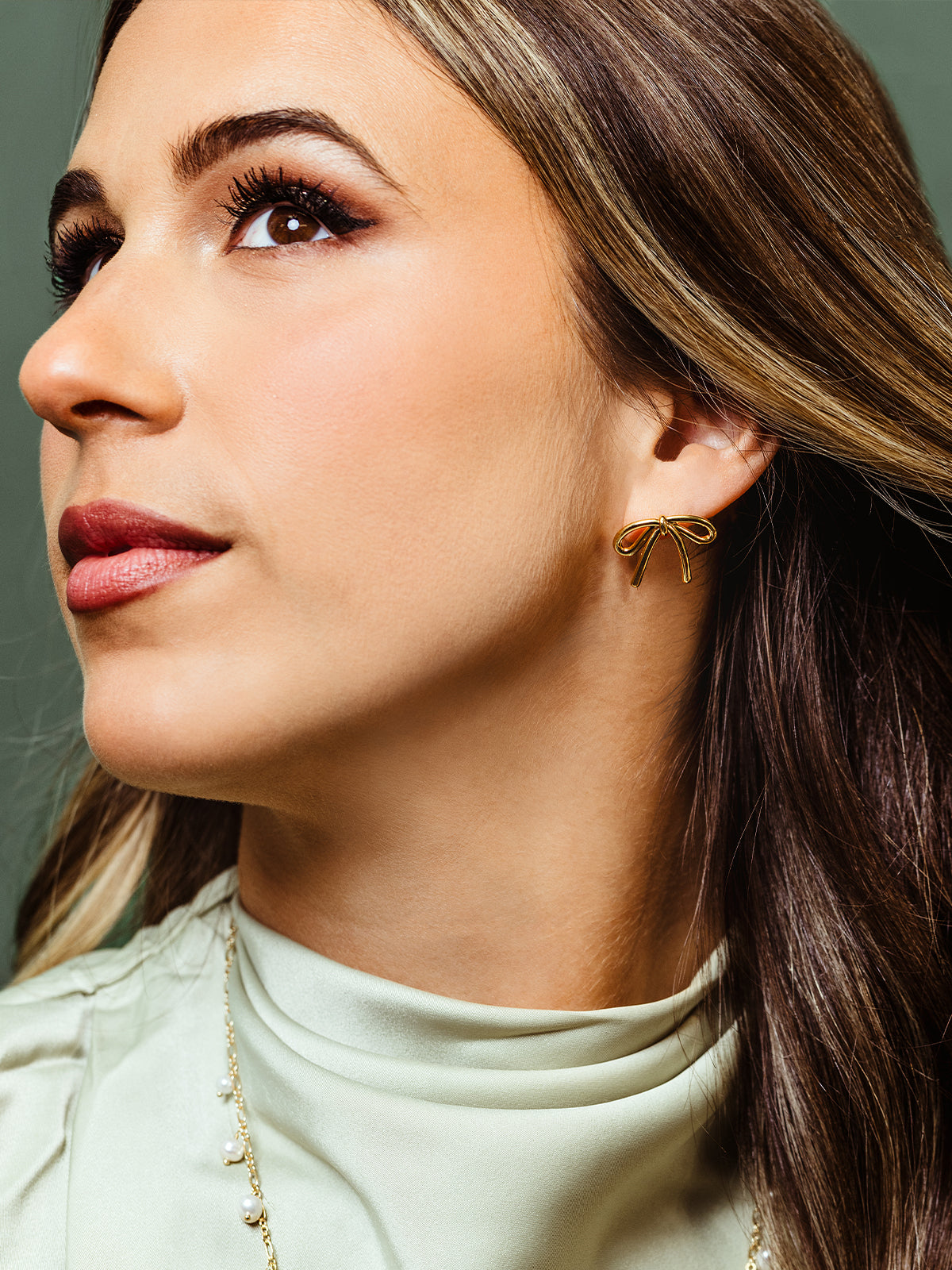 Close-up of a woman wearing delicate gold bow-shaped stud earrings. Her dark hair cascades past her shoulders, and she wears a soft green high-neck blouse, complementing the elegant jewelry.