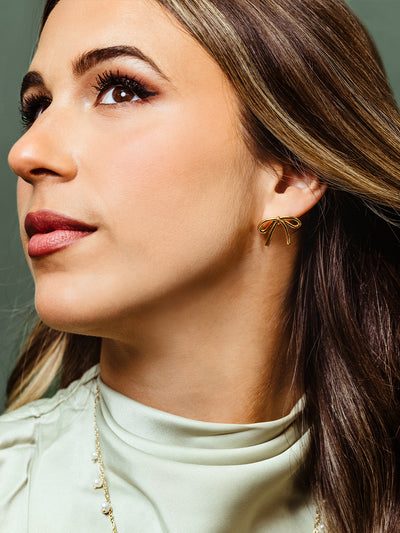 Close-up of a woman wearing delicate gold bow-shaped stud earrings. Her dark hair cascades past her shoulders, and she wears a soft green high-neck blouse, complementing the elegant jewelry.