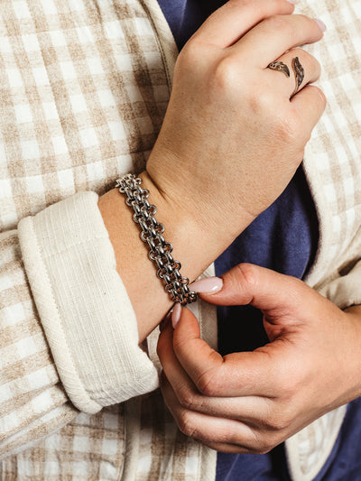 Close-up of a woman's wrist adorned with a silver multi-strand chain bracelet, featuring interwoven links for a bold, textured look. She wears a checkered cream and beige jacket over a navy top, accessorized with a sculptural silver ring, adding a touch of modern elegance.
