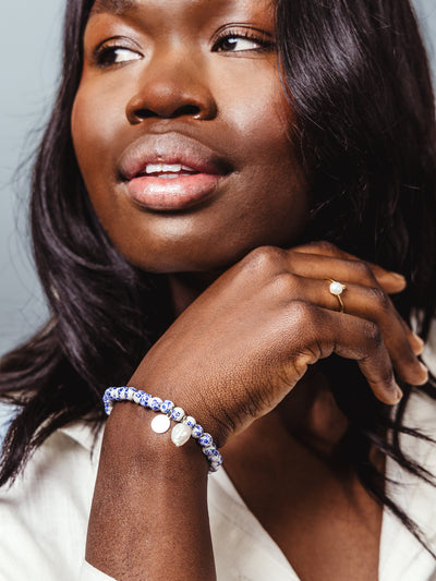 Close-up of a woman wearing a white and blue floral beaded bracelet with pearl and charm accents, styled with a white blouse and gold pearl ring.