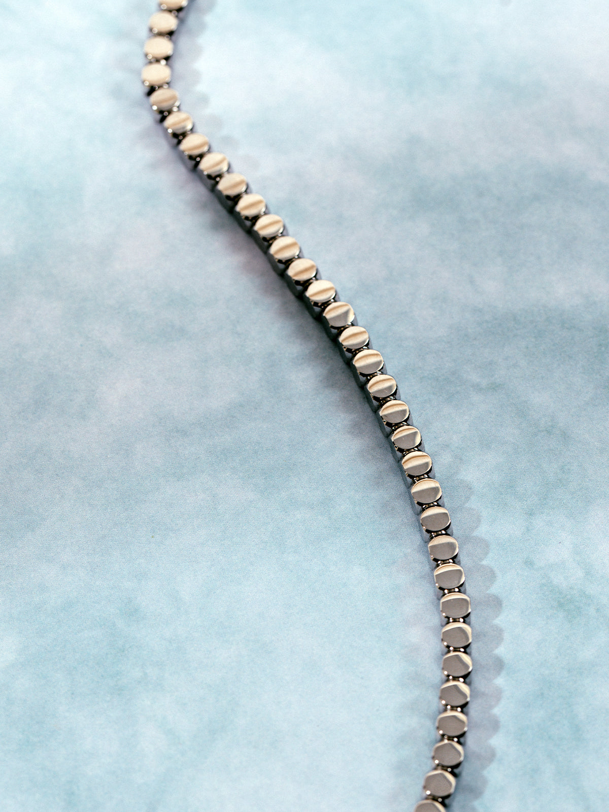 Close-up of a sleek silver-toned chain necklace with geometric circular links, laid out in an S-curve on a soft blue marbled background.