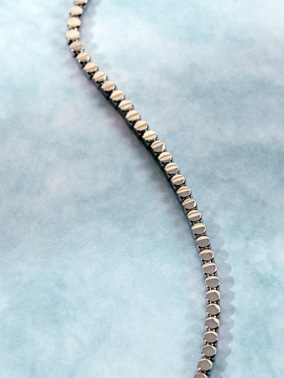 Close-up of a sleek silver-toned chain necklace with geometric circular links, laid out in an S-curve on a soft blue marbled background.