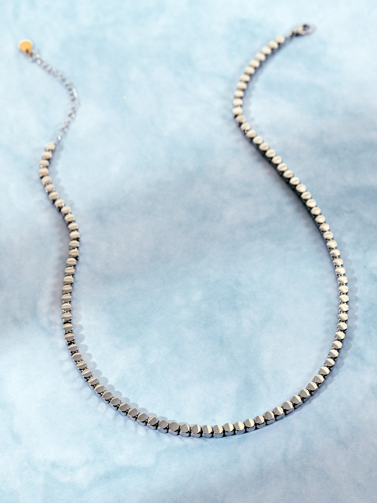 Full view of a silver-toned geometric chain necklace with circular links and extender chain, displayed against a light blue marbled background.