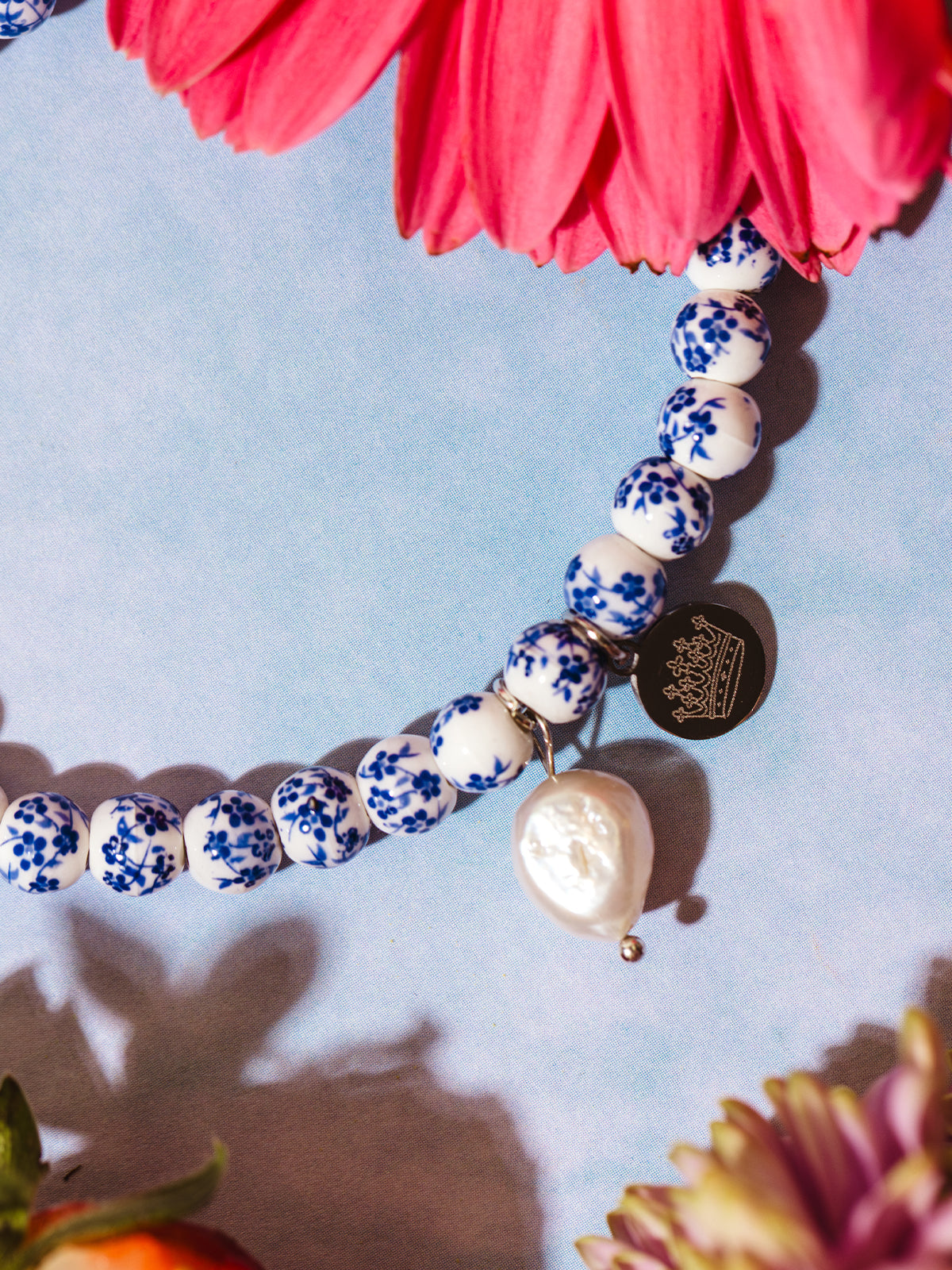 Close-up of a white beaded bracelet with delicate blue floral patterns, featuring a teardrop-shaped pearl charm and a round metallic tag engraved with a crown, surrounded by pink and purple flowers.