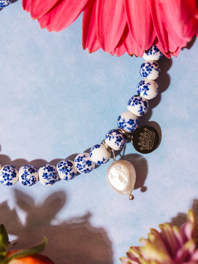 Close-up of a white beaded bracelet with delicate blue floral patterns, featuring a teardrop-shaped pearl charm and a round metallic tag engraved with a crown, surrounded by pink and purple flowers.