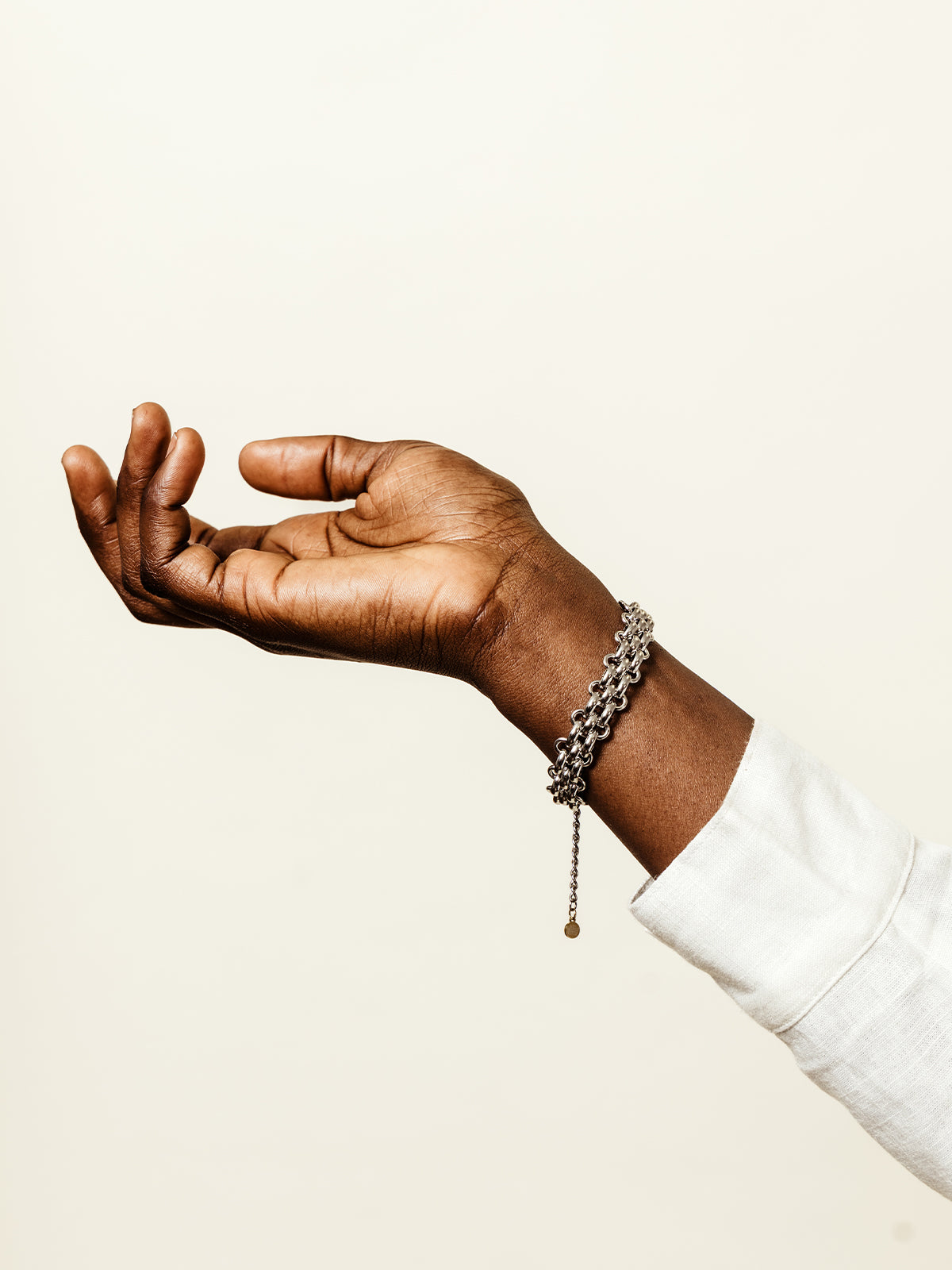 Outstretched hand wearing a silver chain-link bracelet with dangling detail, styled with a long-sleeve white top against a light cream background.