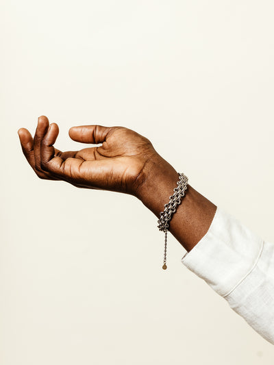 Outstretched hand wearing a silver chain-link bracelet with dangling detail, styled with a long-sleeve white top against a light cream background.