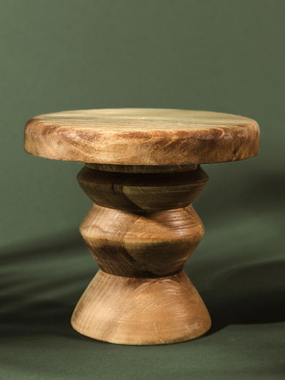 Hand-carved pedestal stand made from rich, dark natural wood with a geometric, sculptural base, set against a forest green backdrop.