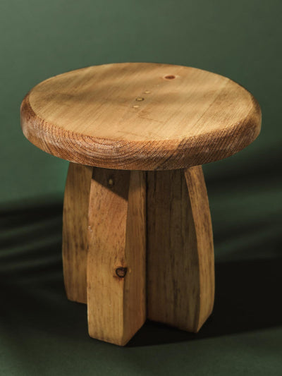 Rustic pedestal stand crafted from dark natural wood, featuring a smooth round top and sturdy vertical legs with organic knots and markings, set against a forest green background.
