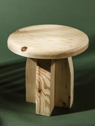 Light natural wood pedestal stand with a round top and clean, geometric legs showcasing wood grain and natural knots, photographed on a dark green surface.